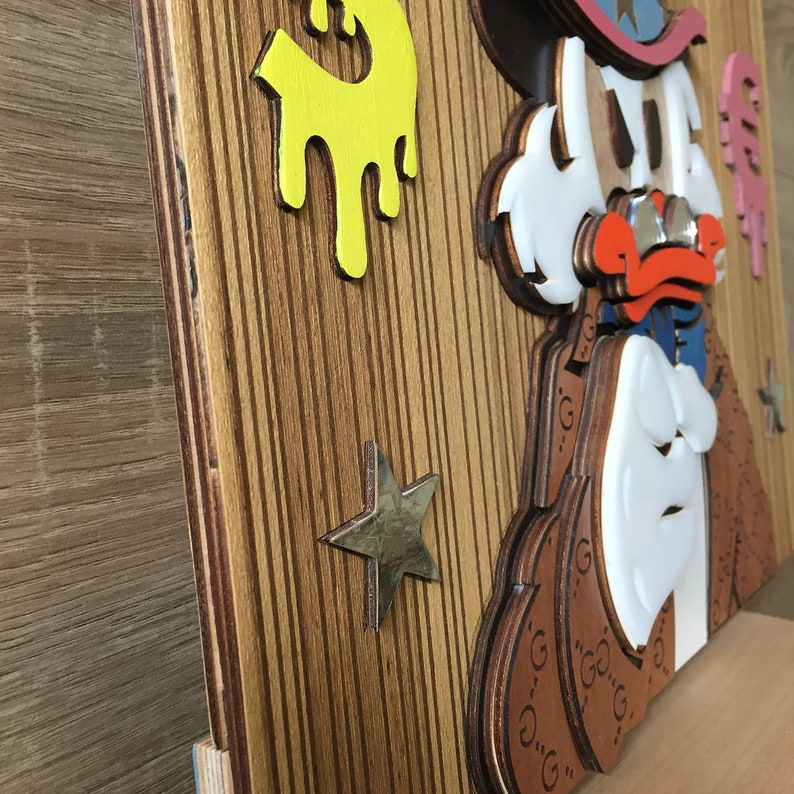 May include: A wooden wall art piece featuring a cartoon character with a cowboy hat, a red nose, and a white mustache. The character is wearing a brown jacket with a pattern of the letter "G". The background is a wood grain pattern with a yellow splatter paint design and a gold star.