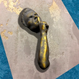 May include: A gold and grey sculpture of a person with their finger to their lips, as if to say "shhh". The sculpture is mounted on a textured, grey wall.