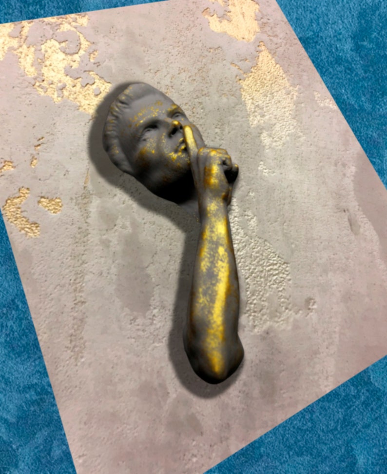May include: A gold and grey sculpture of a person with their finger to their lips, as if to say "shhh". The sculpture is mounted on a textured, grey wall.