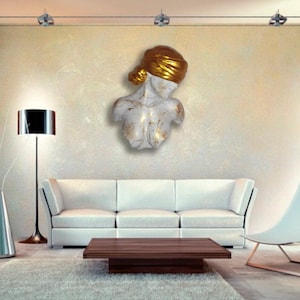 May include: White and gold sculpture of a woman's bust with a gold head scarf. The sculpture is mounted on a wall in a modern living room setting.
