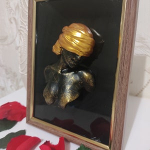May include: A framed 3D wall sculpture of a woman's bust with a gold head covering. The sculpture is set against a black background and framed in a dark wood frame.