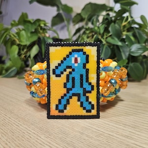 May include: A colorful beaded bracelet with a pixelated image of Squidward from Spongebob Squarepants. The image is on a yellow background with a black border. The bracelet features orange, blue, and yellow beads.
