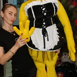May include: A large, yellow plush figure resembling a cartoon character, wearing a black and white maid outfit with a red beaded necklace. The figure has large yellow hands and feet.
