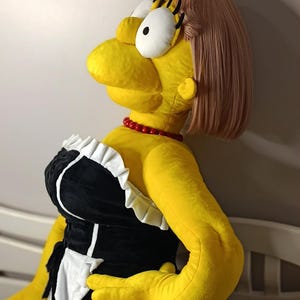 May include: A large plush doll of a cartoon character, dressed in a black and white maid outfit with a red necklace. The doll has yellow skin, a pink wig, and large eyes.