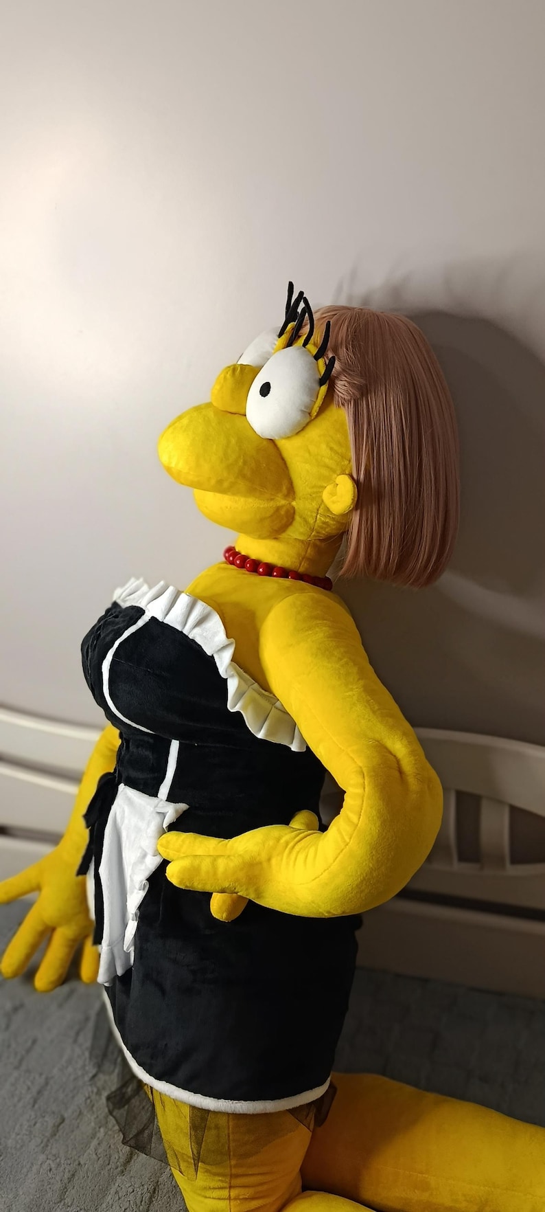 May include: A large plush doll of a cartoon character, dressed in a black and white maid outfit with a red necklace. The doll has yellow skin, a pink wig, and large eyes.