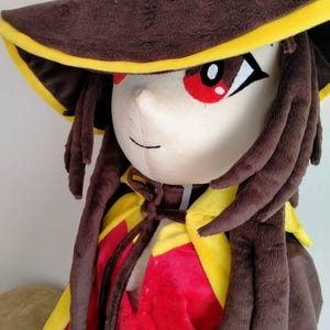 May include: A plush doll of a character wearing a brown hat with yellow trim, a red and yellow cape, and brown hair. The doll has large red eyes and a neutral expression. The doll is made of soft, plush fabric.