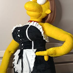 May include: A large plush doll of a cartoon character in a black and white maid outfit with a white apron. The doll has yellow skin, a red necklace, and a blonde wig. The doll is lying down.
