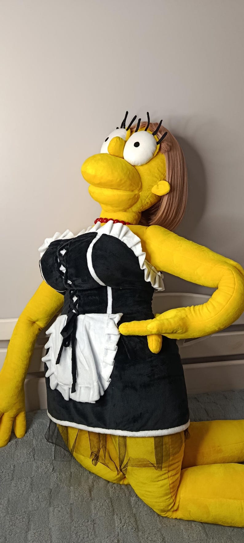 May include: A large plush doll of a cartoon character in a black and white maid outfit with a white apron. The doll has yellow skin, a red necklace, and a blonde wig. The doll is lying down.