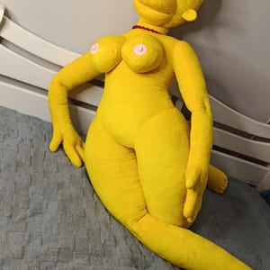 May include: A large, yellow plush doll resembling a cartoon character, with black eyelashes and a red necklace. The doll is lying on a gray textured surface, with its arms and legs extended.