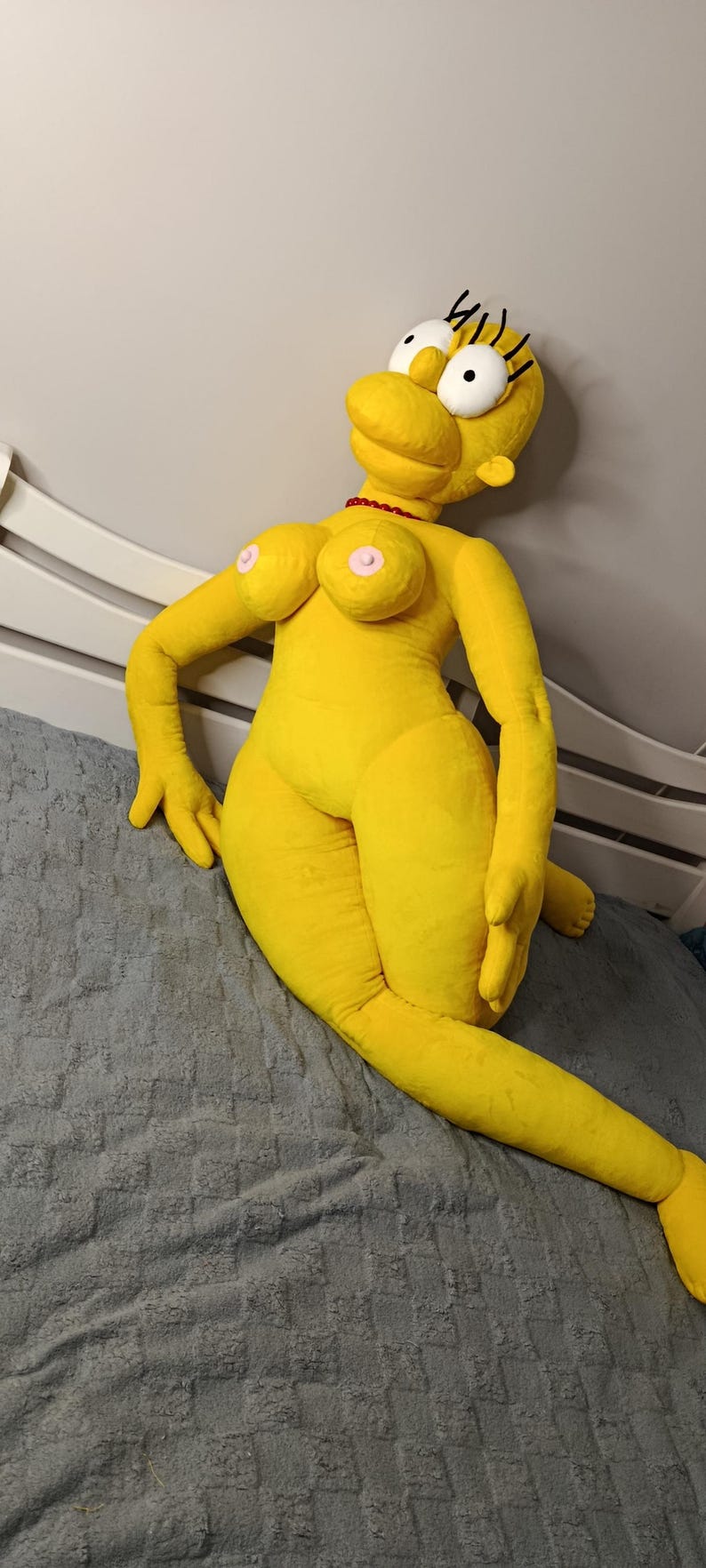 May include: A large, yellow plush doll resembling a cartoon character, with black eyelashes and a red necklace. The doll is lying on a gray textured surface, with its arms and legs extended.