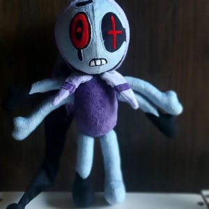 Op de afbeelding: Een pluche pop met een lichtblauwe kop en lichaam, met rode oogdetails en een zwart kruis. De pop heeft een paarse torso, lichtblauwe armen en zwarte accenten. Het heeft een lange, zwarte, puntige staart met een zwarte bal aan het einde.