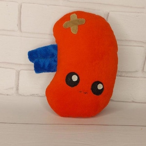 May include: A plush toy shaped like a kidney with a bandage on it. The kidney is orange and has a blue wing on one side. It has black eyes and a small smile.