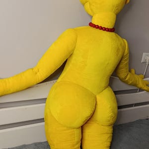 May include: A large, yellow plush doll of a cartoon character, kneeling with arms outstretched. The doll has a red bead necklace and black hair. The doll is on a gray surface.