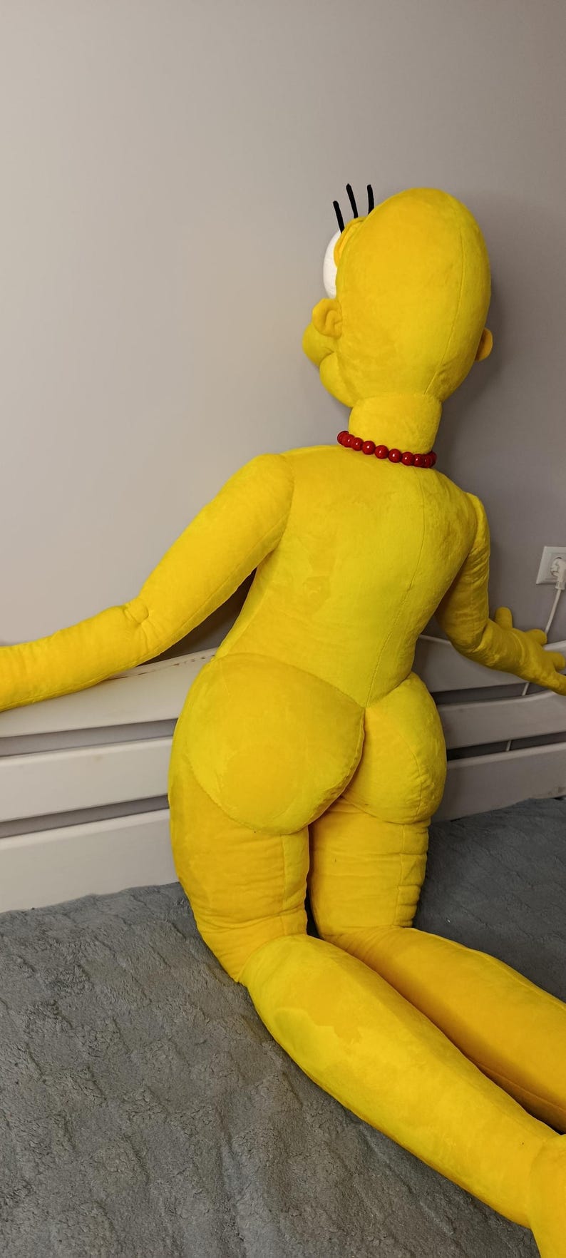 May include: A large, yellow plush doll of a cartoon character, kneeling with arms outstretched. The doll has a red bead necklace and black hair. The doll is on a gray surface.