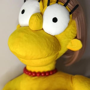 May include: A plush doll of Marge Simpson, featuring her signature yellow skin, blue hair, and a red bead necklace. The doll has large white eyes with black pupils and black eyelashes. She wears a black and white dress.
