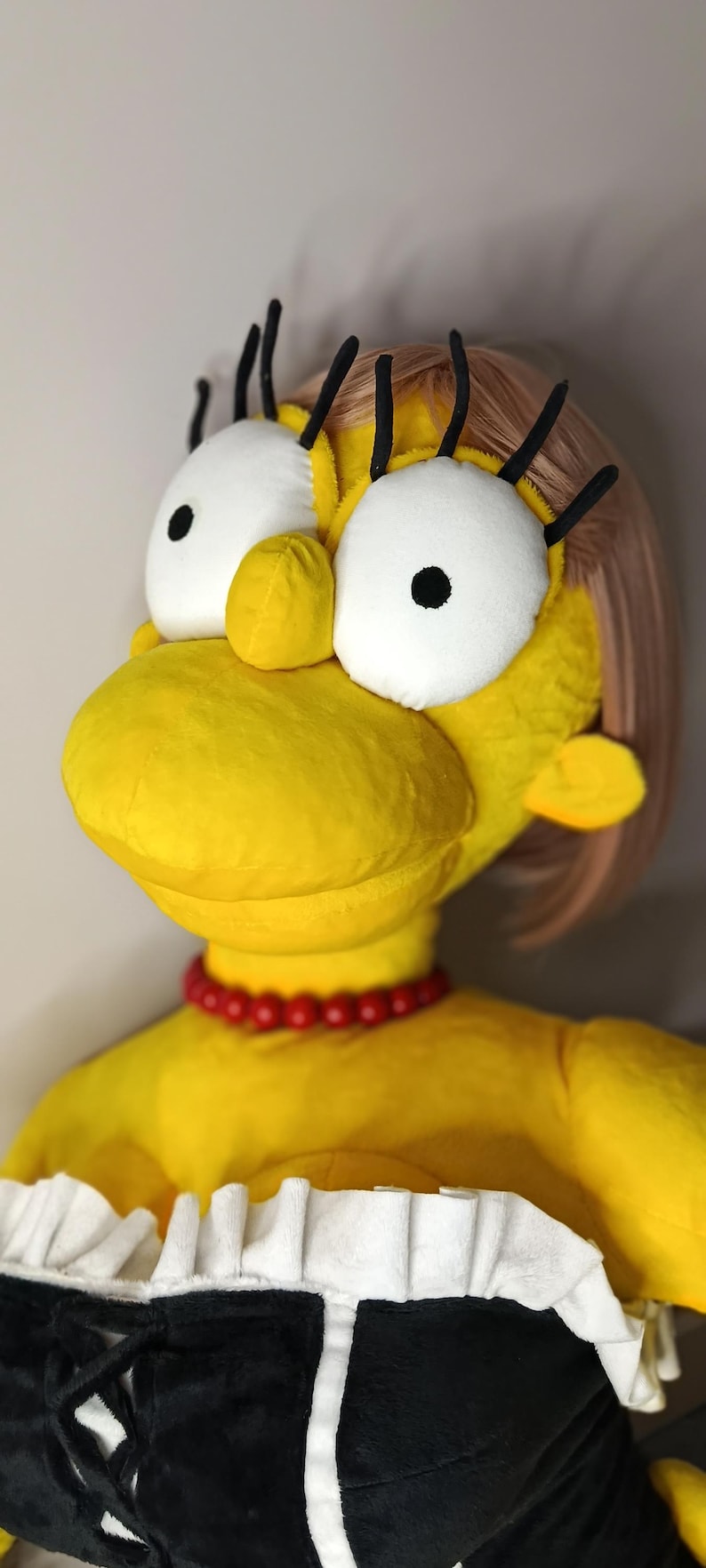 May include: A plush doll of Marge Simpson, featuring her signature yellow skin, blue hair, and a red bead necklace. The doll has large white eyes with black pupils and black eyelashes. She wears a black and white dress.