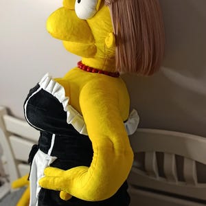 May include: A large plush doll of a cartoon character in a maid outfit. The doll is yellow with a pink wig, a red bead necklace, and a black and white dress. The doll is kneeling on a gray surface.