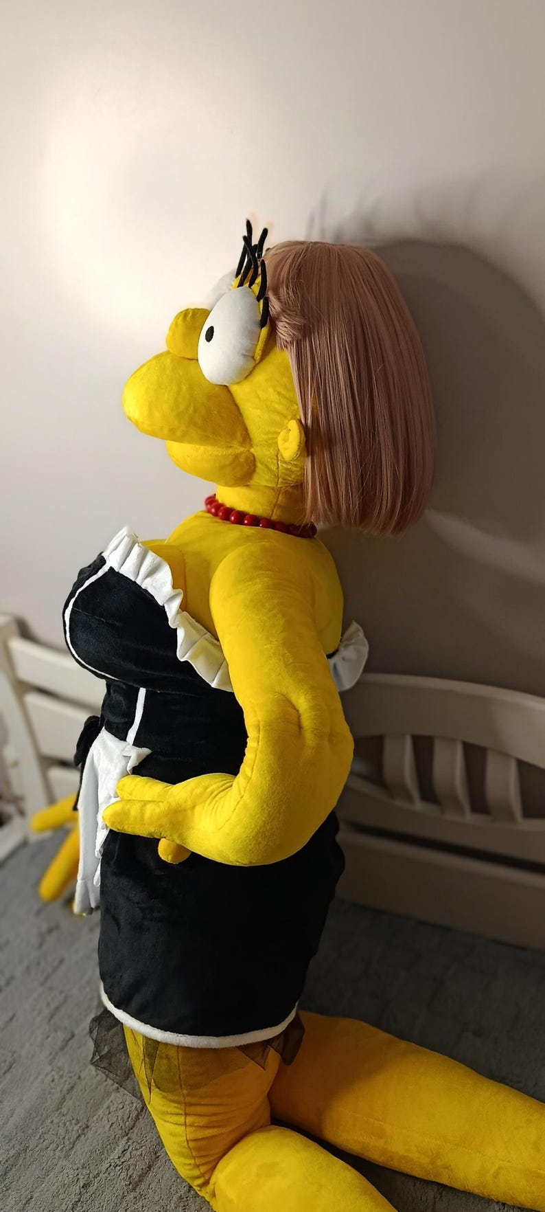 May include: A large plush doll of a cartoon character in a maid outfit. The doll is yellow with a pink wig, a red bead necklace, and a black and white dress. The doll is kneeling on a gray surface.