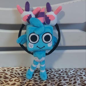May include: A light blue plush toy character with a square head, large black eyes, and a smiling mouth. The toy has small blue arms and legs, and wears a diaper. Two white hands with pink and purple accents are attached to a black headband.