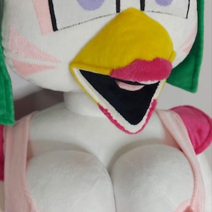 Op de afbeelding: Een pluche speelgoed van een cartoon kippenkarakter met witte, roze, gele en zwarte accenten. Het karakter heeft grote ogen, een gele snavel en een roze top. Het speelgoed is zacht en knuffelbaar.