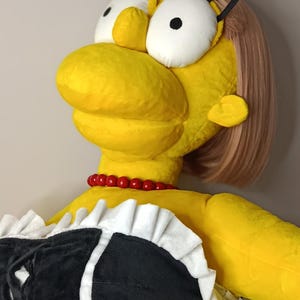 May include: A large plush doll of a cartoon character with yellow skin, a red bead necklace, and a black and white outfit. The doll has large white eyes, black eyelashes, and a pink hairstyle.