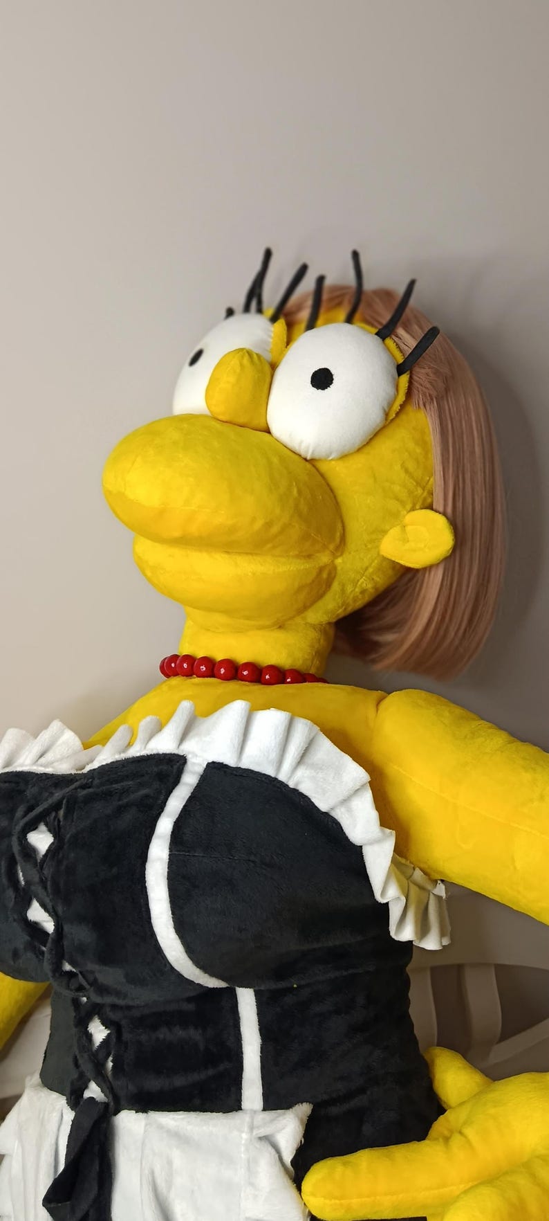 May include: A large plush doll of a cartoon character with yellow skin, a red bead necklace, and a black and white outfit. The doll has large white eyes, black eyelashes, and a pink hairstyle.