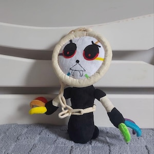 Puede incluir: Muñeca de peluche hecha a mano con una cabeza circular con ojos rojos y una boca cosida. La muñeca tiene un cuerpo negro, cabeza y brazos color crema y manos de colores del arcoíris. Una cadena está envuelta alrededor del torso de la muñeca.