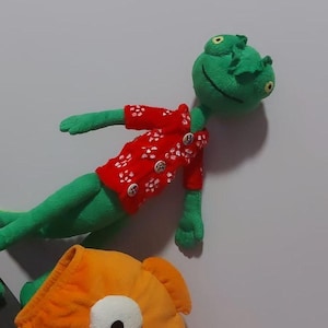 Pode incluir: Um sapo de pelúcia verde vestindo uma camisa vermelha de manga curta com um padrão floral branco e quatro botões, posicionado acima de um grande peixe de pelúcia laranja com um olho branco proeminente e uma pupila preta.