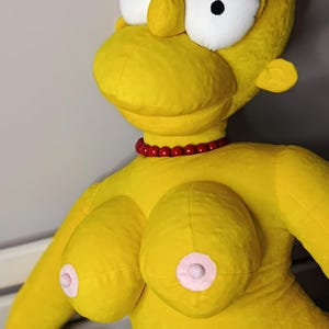 May include: A large, plush yellow doll resembling a cartoon character. It features oversized white eyes with black pupils, black eyelashes, and a red beaded necklace. The doll has large breasts with pink nipples.