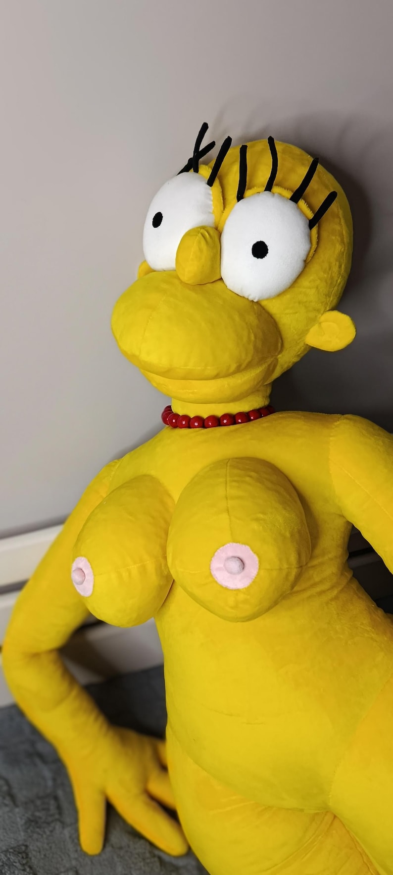 May include: A large, plush yellow doll resembling a cartoon character. It features oversized white eyes with black pupils, black eyelashes, and a red beaded necklace. The doll has large breasts with pink nipples.