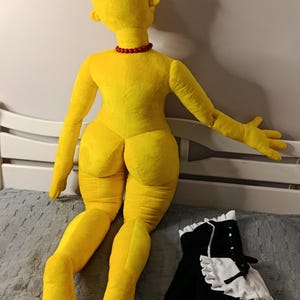 May include: A large, yellow plush doll with a red necklace and a black and white maid outfit. The doll is lying on a gray surface. The doll's features include a round head, large feet, and a distinctive hairstyle.