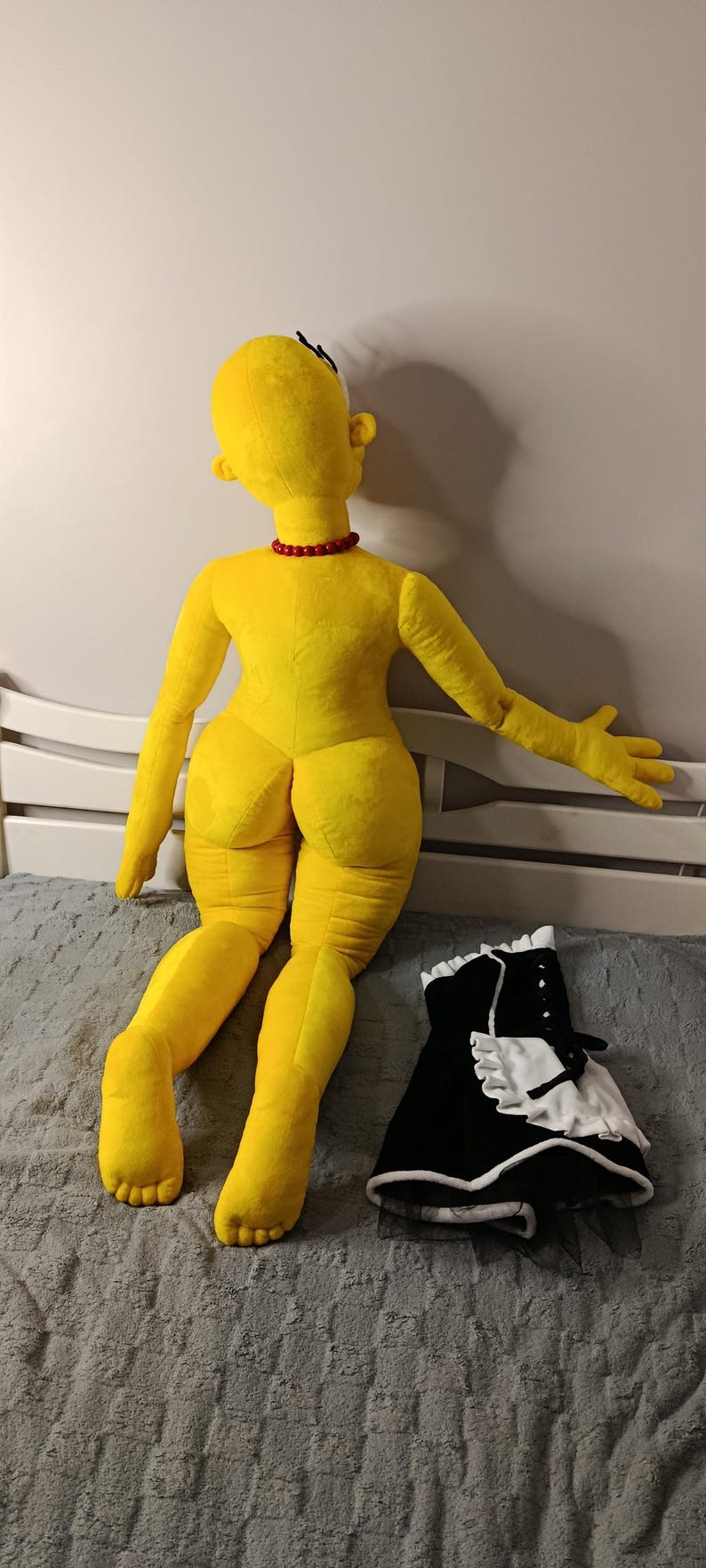 May include: A large, yellow plush doll with a red necklace and a black and white maid outfit. The doll is lying on a gray surface. The doll's features include a round head, large feet, and a distinctive hairstyle.