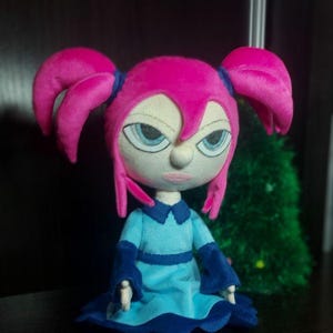 May include: A handmade doll with a stylized design. The doll features a pink head with two large, curved pigtails, blue eyes, and a light blue dress with dark blue accents. The doll's expression appears stern.