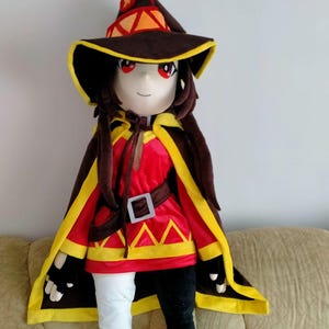 May include: A plush doll dressed as a character from an anime series. The doll wears a red and yellow outfit with a brown hat and cape. The doll has red eyes and long brown hair. The doll is approximately 36 inches tall.