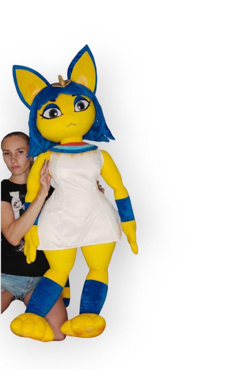 May include: A large yellow plush toy with blue hair and ears, wearing a white dress and blue leg warmers. The toy is standing next to a person.