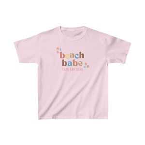 Peut inclure: T-shirt rose avec le texte "beach babe" en lettres colorées et un petit graphique d'une coquille, d'une étoile de mer et d'une vague. Le texte "CAPE SAN BLAS" est imprimé sous le texte principal.