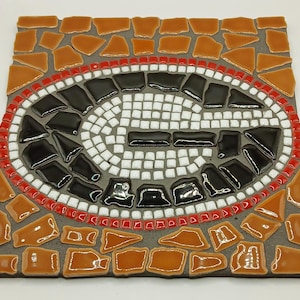 May include: A square mosaic tile featuring a large white letter "G" in the center, surrounded by black tiles and a red border. The outer border is made of orange and brown tiles, creating a decorative piece.