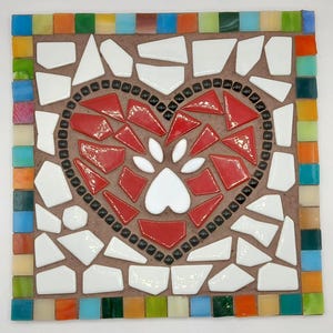 May include: Square mosaic art piece featuring a heart design with a paw print. The heart is red, surrounded by black and white tiles. The border is a mix of colorful tiles, including orange, green, and blue. The background is brown.