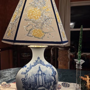 Może przedstawiać: Dekoracyjna lampa stołowa z beżowym abażurem z żółtymi i srebrnymi kwiatowymi wzorami i granatowym wykończeniem. Podstawa lampy to biały porcelanowy wazon z niebieskimi ilustracjami krajobrazowymi.