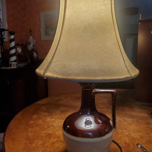 May include: A brown and white ceramic lamp base shaped like a jug with a handle. The lamp has a beige fabric lampshade.