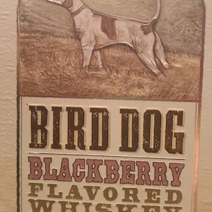 Op de afbeelding: Een illustratie van een bruine en witte vogelhond op een witte achtergrond met de tekst "Bird Dog Blackberry Flavored Whiskey" in gouden en bruine letters. De fles is 80 proof en 40% alc./vol. 1.75L.