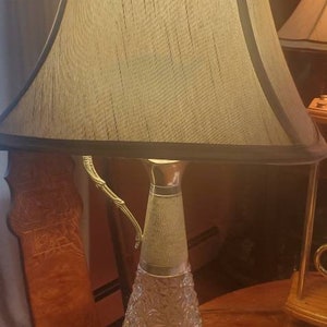 Może przedstawiać: Lampa stołowa w stylu vintage z przezroczystą szklaną podstawą i złotym akcentem metalowym. Lampa ma beżowy abażur z czarną lamówką.