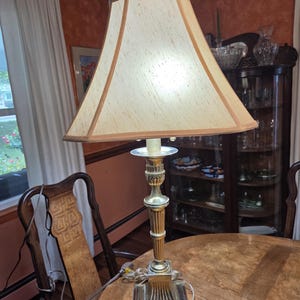 May include: A table lamp with a metallic gold-toned base featuring a square pedestal and a fluted column. The lamp is topped with a beige, four-sided, tapered fabric lampshade with matching trim along the edges and a small finial at the top.