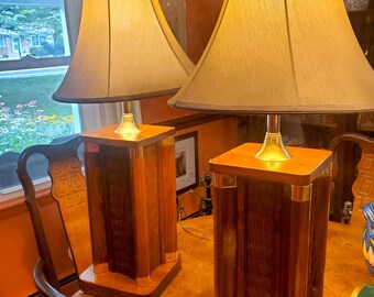 May include: Two matching table lamps with tan shades and wooden bases. The bases are rectangular with a dark wood finish and brass accents. The lamps are placed on a wooden table, creating a warm and inviting ambiance.