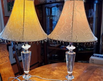 Puede incluir: Dos lámparas de mesa de cristal con pantallas beige con un patrón de espiral. Las pantallas tienen un ribete de flecos con cuentas.