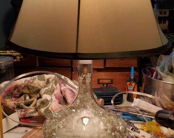 May include: A table lamp with a beige lampshade and a clear glass base filled with small, clear crystals. The lamp is on a white table with a wooden cutting board in front of it.