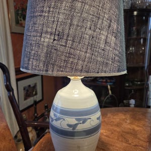 May include: A ceramic table lamp with a white base and blue leaf design. The lamp has a navy blue and white textured lampshade. The lamp is sitting on a wooden table.