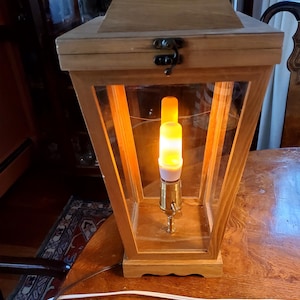 以下が含まれることがあります： ガラス製の前面と、暖色系の白色LEDキャンドルが内蔵された木製のランタン。ランタンは木製のテーブルの上に置かれています。