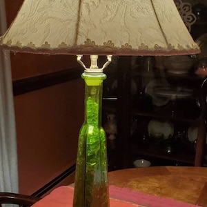 May include: A green glass bottle lamp with a white fabric lampshade with a floral pattern. The lamp is on a table with a red and orange patterned runner.