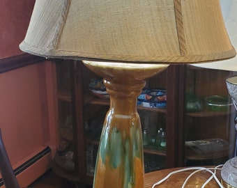 May include: A ceramic table lamp with a brown and green glazed base and a beige fabric lampshade. The lampshade has a brown trim around the top and bottom.